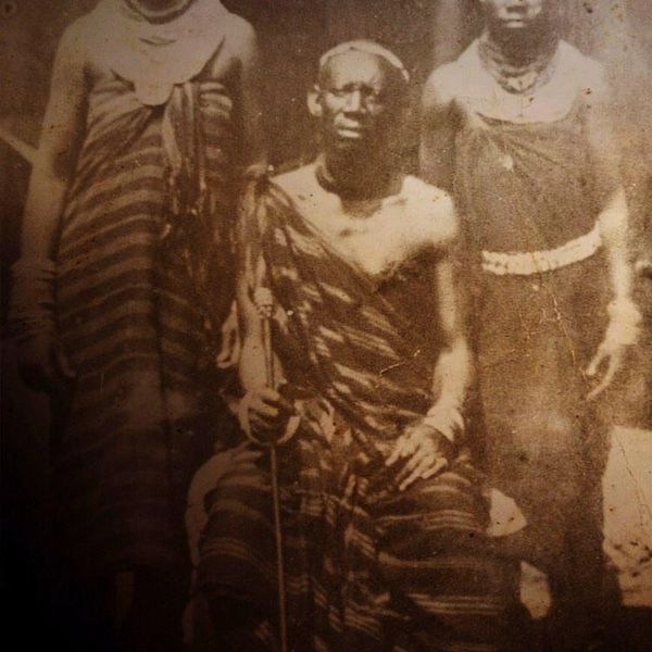 Old sepia photo of three people in traditional attire, one seated with a staff.