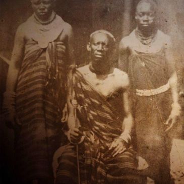 Old sepia photo of three people in traditional attire, one seated with a staff.