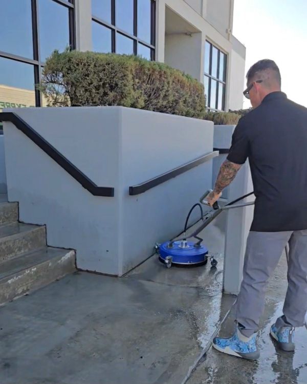 Man cleaning sidewalk with a blue surface cleaner near stairs.