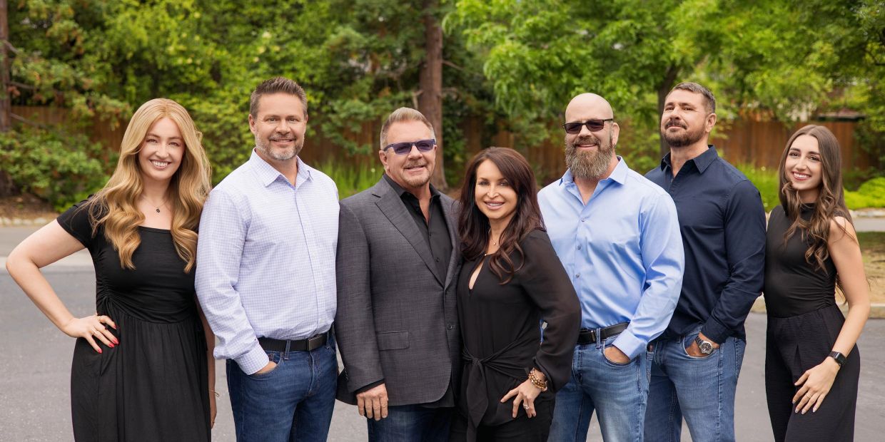 Seven smiling adults posing outdoors together.