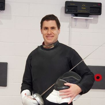 Smiling fencer in protective gear holding helmet and foil indoors.