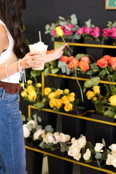 Woman picking a yellow rose while holding an iced coffee in a flower shop.