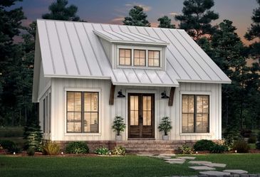 Modern small white house with metal roof and warm lit windows at dusk.