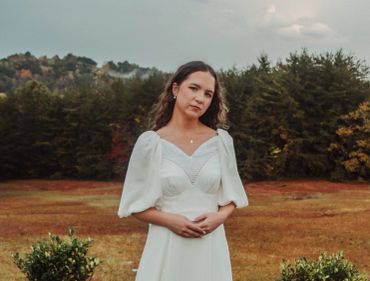 A woman in a white dress standing in a field with trees in the background.