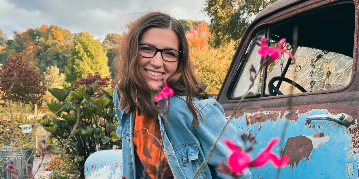 Smiling woman in glasses poses by an old rusty blue truck with autumn foliage in background.