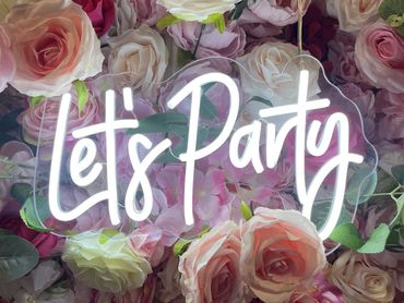 Neon sign saying 'Let's Party' on a floral backdrop.
