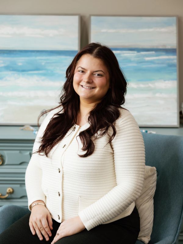 Smiling woman with dark hair in a white sweater sitting on a blue chair.