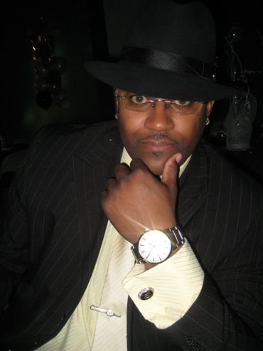 Man in pinstripe suit and fedora posing thoughtfully with watch visible.