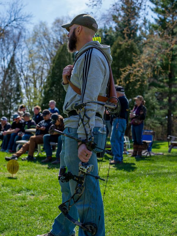 Man in casual attire holding a compound bow outdoors with spectators in the background.
