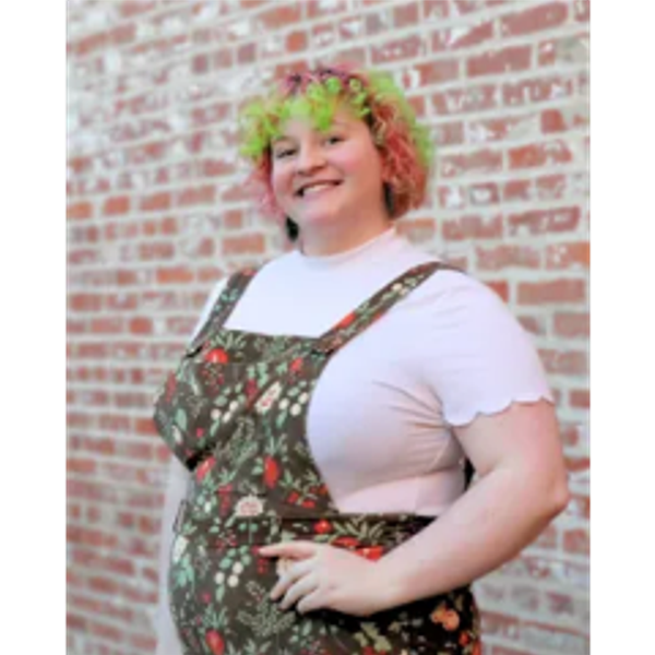 Smiling person with curly, colorful hair in floral overalls against a brick wall.