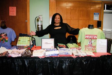 Woman smiling behind a table displaying various colorful T-shirts for sale.
