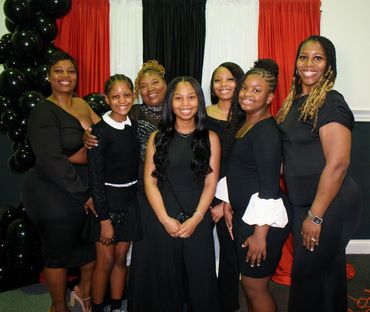 A group of seven women dressed elegantly in black poses smiling in front of red, white, and black drapes.