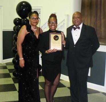 Three people dressed formally at an award ceremony, one holding a plaque.