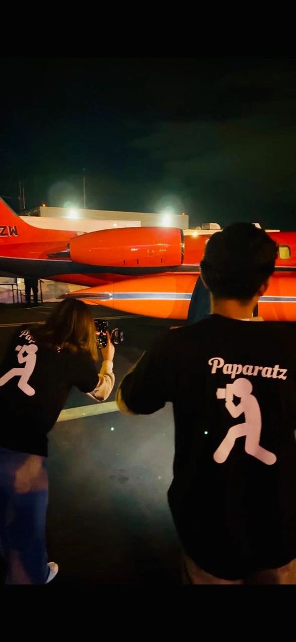 Two photographers in 'Paparatz' shirts capturing a red airplane at night.