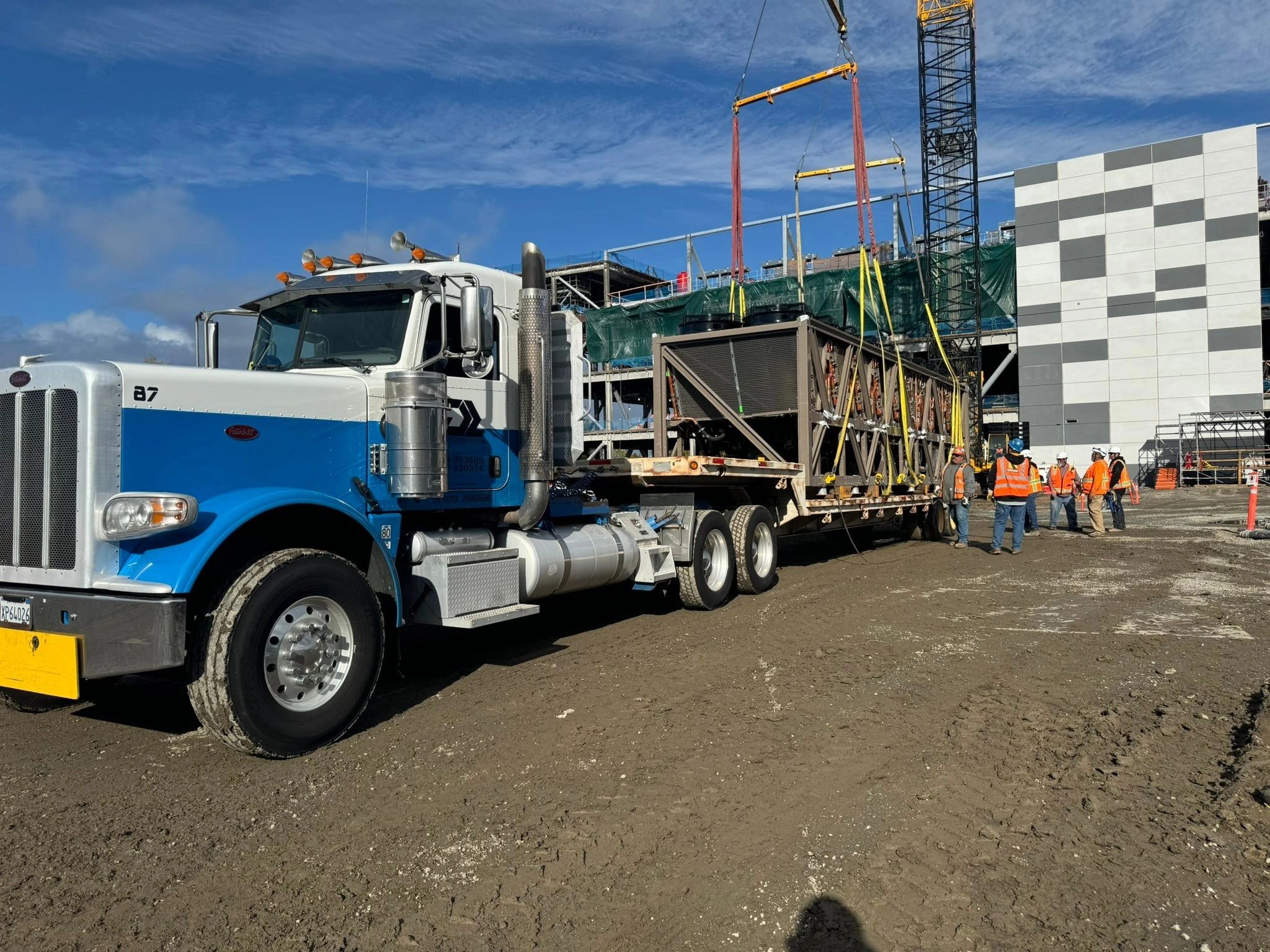 Construction workers guide a crane lifting equipment from a blue and white truck.