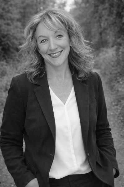 Smiling woman in a navy blazer stands outdoors on a wooded path.