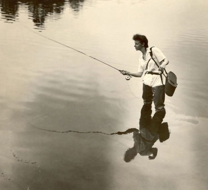 Joan Wulff standing in still water with a fly fishing bag over her shoulder and a fly rod in her han
