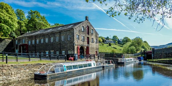 Historic stone building by a calm canal with moored boats under a bright blue sky.