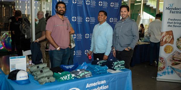 Three men stand behind a table with American Foundation merchandise at an event.