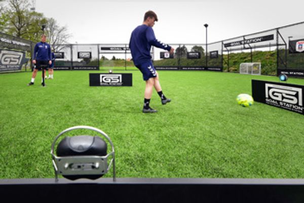 Two men playing soccer on a small artificial turf field with goal station branding.