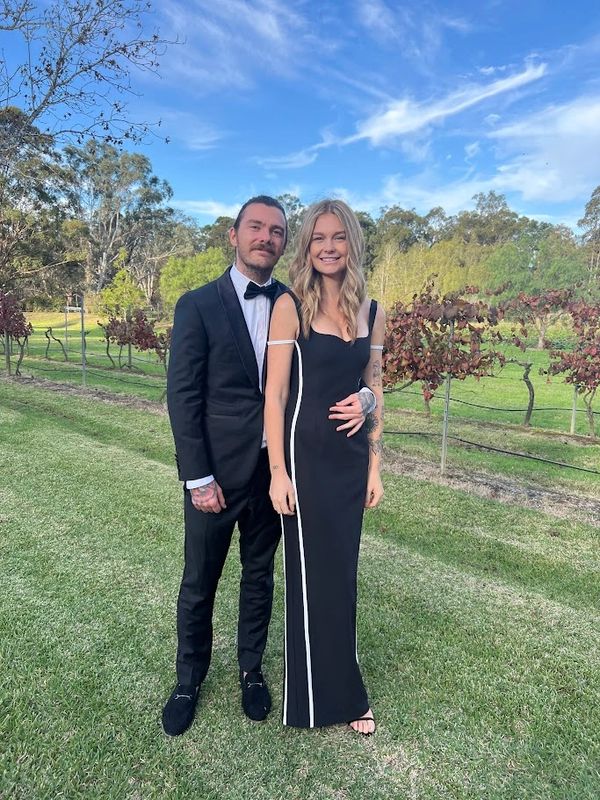 A couple dressed elegantly in black stands outdoors on lush green grass under a blue sky.