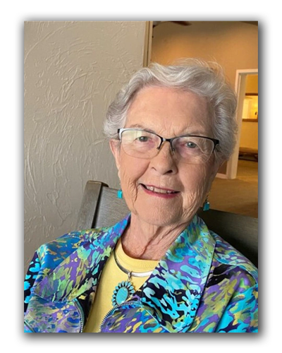 Smiling elderly woman with glasses wearing a colorful jacket and turquoise jewelry.