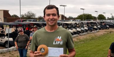 Man smiling, holding a trophy and a check at a golf course.