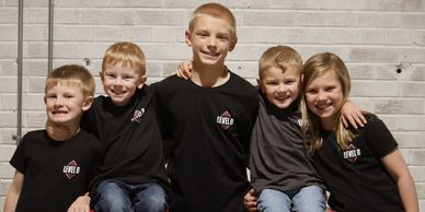 Five children smiling and posing together against a brick wall.