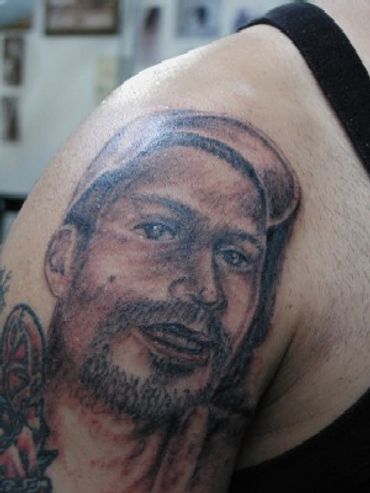 Black and grey portrait tattoo of a man with a baseball cap on a shoulder.