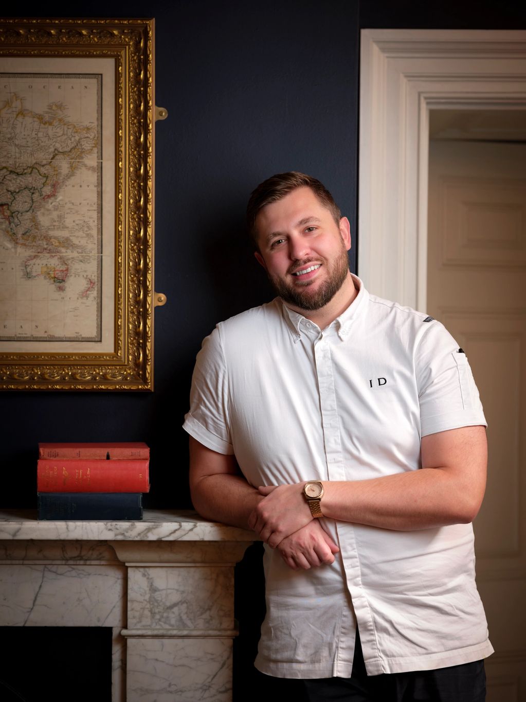 Smiling man leaning against a marble fireplace with books and a framed map.