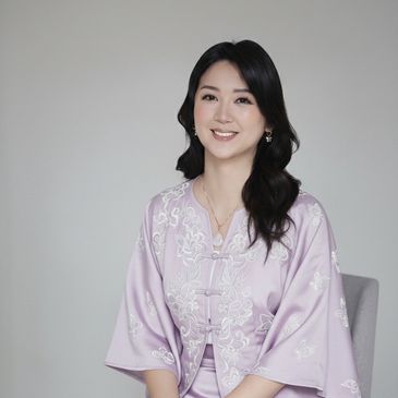 Smiling woman in a lavender embroidered traditional outfit sitting on a chair.