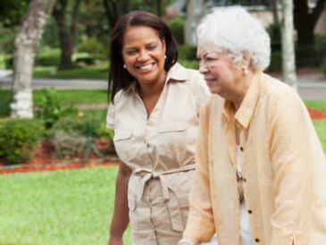 A younger woman assists an elderly woman using a walker outdoors.