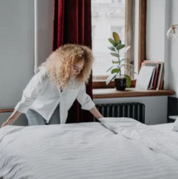 A person making a bed in a cozy, well-lit bedroom.