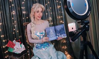 Woman dressed as Elsa from Frozen reading a book on camera with festive lights.