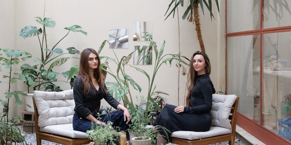 Two women seated in a cozy indoor garden with plants and wooden furniture.