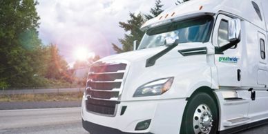 White Freightliner truck driving on a highway with trees and sun in background.