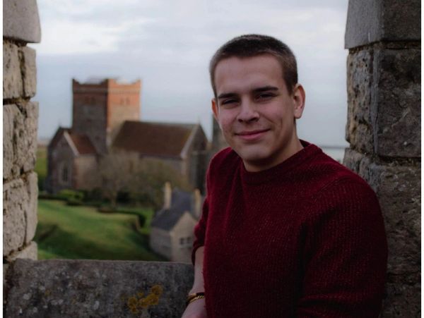 A young man leaning on a stone wall with a blurred castle in the background.
