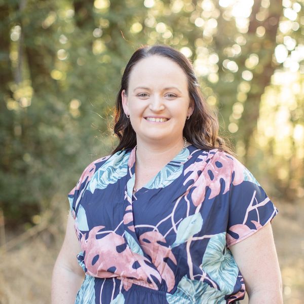 Smiling woman in a floral dress standing outdoors in soft sunlight.