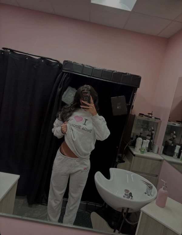 Person in gray sweatpants and sweatshirt taking a mirror selfie in a salon.