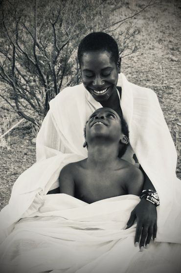 Desert Queen and son wrapped in white fabric seated on desert ground. Smiling at each other.