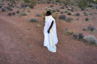 Aerial drone photo side view of desert woman draped in white fabric with mountains and clouds.