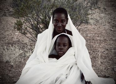 Desert Queen and son wrapped in white fabric seated on desert ground.