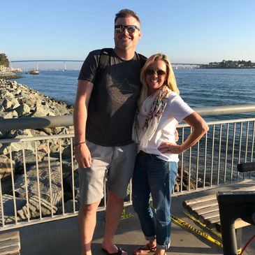 A smiling couple posing by the waterfront with a bridge in the background on a sunny day.