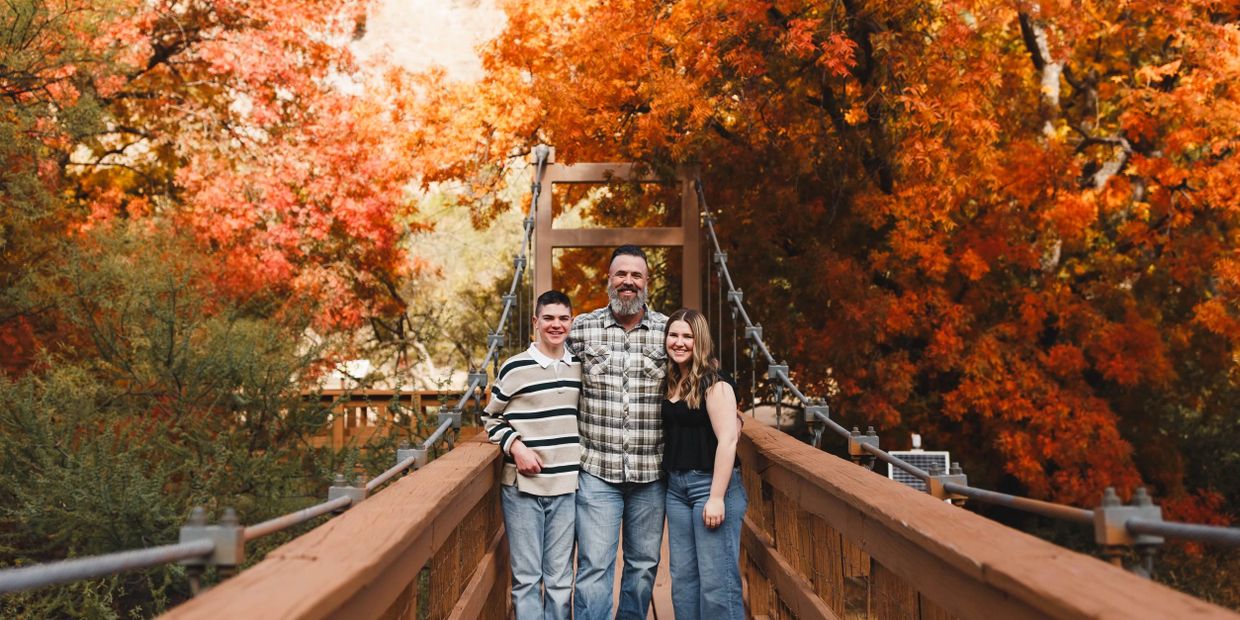 Family of three smiling on a wooden bridge surrounded by vibrant autumn trees.