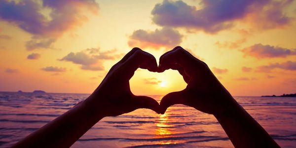 Hands forming a heart shape at sunset over the ocean.