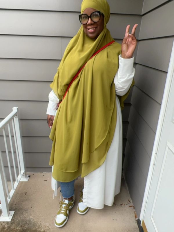 Woman in olive green hijab smiling and making a peace sign.