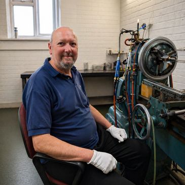 Man in blue shirt and white gloves sitting by industrial machinery.