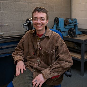 A smiling young man sitting in a workshop with machinery in the background.