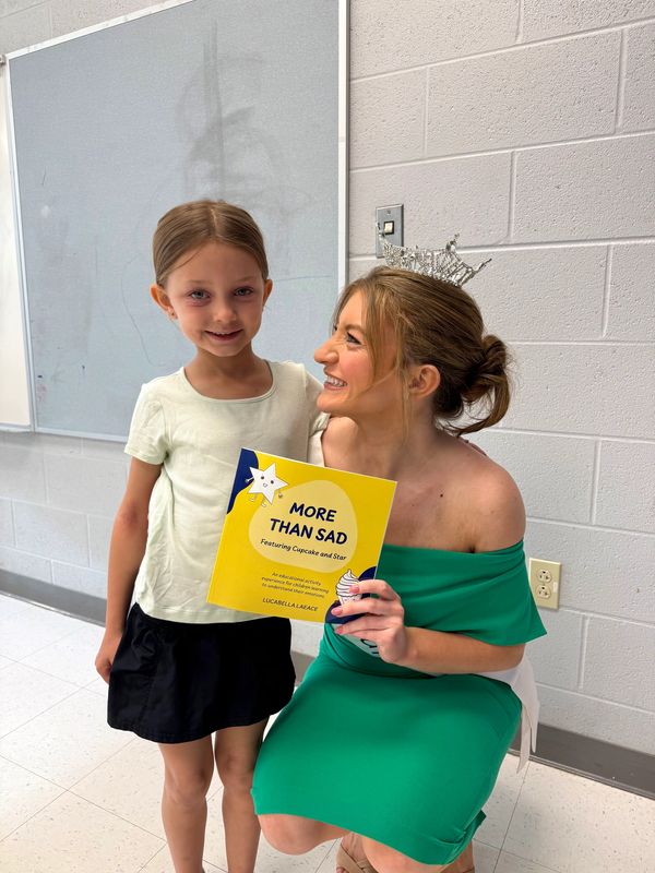 A woman in a green dress and crown smiles at a young girl holding a book.