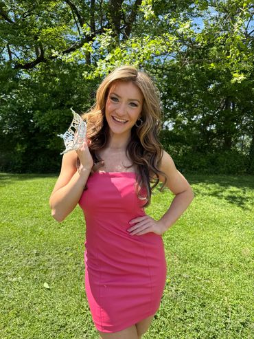 Young woman in a pink dress holding a crown, standing outdoors on grass.